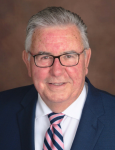 A smiling white male with greying hair wearing glasses, a striped blue and navy blue tie, white shirt, and navy blue jacket.