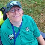 Norman A. Smith looking up at camera smiling dress in a green shirt and cap with CERT written on both