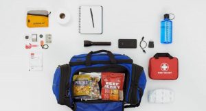 Various items are shown including a flashlight, first aid kit, water bottle, notepad and pen, packaged food in a duffle bag