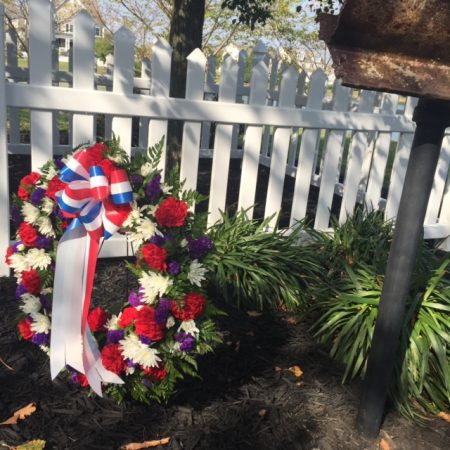 A wreath f red, white, and blue flowers stand before a white picket fence and next to a pol with charred piece of steel