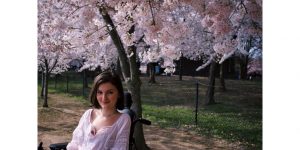 A young white woman with chin-length brown hair smiles at the camera. She wears a white shirt and sits in a black motorized wheelchair. Behind her are several blooming cherry blossom trees.