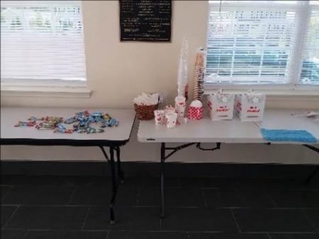 Two white rectangle tables with coffee in disposable boxes, donuts, and breakfast bars.