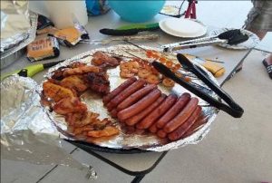 Hotsdogs and other grilled food items on a serving plate with servings tongs. Two hamburgers are on the table with other food.